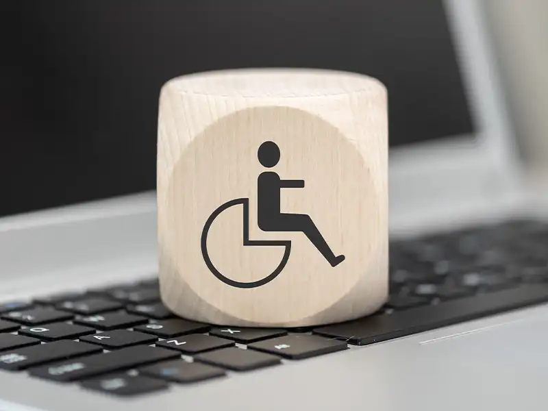 Ein Holzwürfel mit einem Icon darauf steht auf einer Tastatur. Das Icon auf dem Würfel zeigt eine Person in einem Rollstuhl. Es geht um digitale Barrierefreiheit.